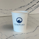 A 10 oz. white paper cup with a blue Coastal Cabinet Works logo featuring a wave, placed on a marble surface. Printed by The Cup Store.