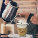 A person pours hot water from a stainless steel kettle into a 10 oz white paper coffee cup with a custom coffee sleeve and steam is rising from the cup. Cup and sleeves sold by The Cup Store.