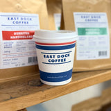 10 oz. Multicolor Recyclable Paper Cup with "east dock coffee" logo, on a wooden shelf, in front of bags of coffee beans