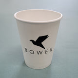 A white 12 oz. custom-printed coffee cup logoed with Bower, sits on a blue surface. The cup is printed by The Cup Store.