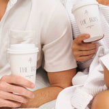 Two people, one in a white shirt and the other in a white bathrobe, are seated close together, holding 12 oz. custom-printed paper cups logoed The Iris Charleston, SC. Only their torsos and hands are visible, each person holding a cup in one hand. The cups are printed by The Cup Store.