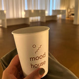 A hand is holding a 12 oz. custom-printed paper cup with the logo mood house. The background features a clean environment and a scenic spa. The cups are printed by The Cup Store.