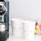 A coffee machine sits on a counter next to two 12 oz. custom-printed paper cups labeled "THE IRIS, CHARLESTON SC." Beside the cups is a clear container holding various colored coffee pods. The cups are printed by The Cup Store.