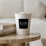 A white 12 oz. custom-printed paper cup with a black logo labeled "ICON Studio Space" sits on a round marble table. The background is blurred, showcasing neutral-toned furniture and a cozy, modern indoor setting. The cups are printed by The Cup Store.