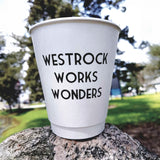 A custom double walled paper coffee cup with WESTROCK WORKS WONDERS printed in bold black letters is placed on top of a large rock. This double-walled cup sits comfortably in a grassy area with trees and a building slightly out of focus, showcasing both premium insulation and custom printing. Printed by The Cup Store
