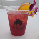 12 oz plastic cup filled with sparkling red drink with ice, garnished with orange and flower, with "Bali Hai" palm logo, on white table