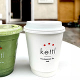 12 oz double-walled paper cup with white dome lid, "Kettl, Fine Japanese Tea" logo with three red dots, next to plastic cup with green smoothie, on white table