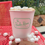 12 oz paper cup with green "The Powder Room" logo, filled with hot chocolate and mini marshmallows, on red metal table next to chocolate pieces and marshmallows