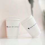 Two 12 oz double-walled paper cups with black "Skin by Lovely" logo and hummingbird, leaning against each other on white table, pastel pink marble wall
