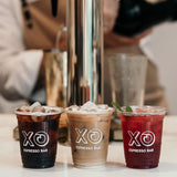 Three 12 oz plastic cups with XO Espresso Bar logo, filled with colorful iced drinks on white table with coffee machine in background