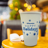 16 oz. Custom Printed Recyclable Paper Cup logoed with Happy Holidays on it, sitting on a cafe table with a book and christmas tree in the background. Cup printed by The Cup Store.