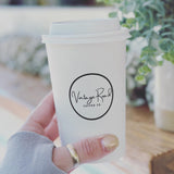Hand holding a 16 oz white coffee cup labeled "Vintage Road Coffee Co." in front of a blurred background with green plants. Cup printed by The Cup Store.