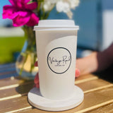 A person holds a 16 oz custom-printed white paper cup with a "Vintage Road Coffee Co." logo on a wooden table. Bright flowers are blurred in the background. Cup printed by The Cup Store.