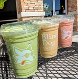 Three drinks - green, cream, and red, in cups with Blind Dog Coffee logo and sip lids, on black metal table, cafe brick wall in background