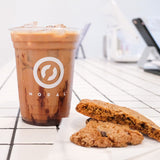 16 oz plastic cup with "Modal" logo, filled with iced coffee and chocolate syrup, on white tiled table next to two cookies in cafe