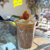 16 oz plastic cup with white logo, filled with iced milk coffee decorated with brown ice teddy bear, on glass table with cafe in background