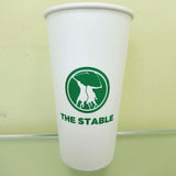 20 oz. Custom Printed Recyclable Paper Cup labeled with The Stable, sits on a glass surface. Cup printed by The Cup Store.