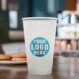20 oz. Custom Printed Recyclable Paper Cup with Your Logo Here, sitting on a coffee table with cookies in the background. The setting is in a cafe with a blurred effect. Cups provided by The Cup Store.