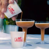 4 oz custom-printed paper cup labeled "Café Aroma" into a cocktail glass. Another cocktail glass with the same brown liquid is already filled and placed nearby on a white tablecloth. One more recyclable "Café Aroma" cup is also visible in front of the cocktail glass. Cups are printed by The Cup Store.