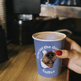8 oz. Blue Multicolor Recyclable Paper Cup with "who let the dog coffee", held on top of a wooden coffee bar counter.
