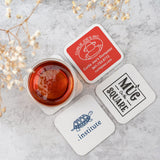 Four square coasters with various logos, with a cup of tea, on a marble table next to a plant