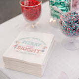 Stack of White Paper Sandwich Bags with "Merry and Bright" print, next to various cups filled with candy, on a white cloth table