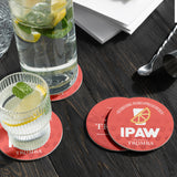Four red round coasters, with white "IPAW" logo and lemon slice image, next to glass and a jug of lemonade, on black wooden table
