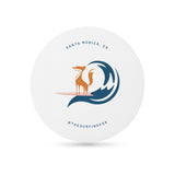 White round coaster with an illustration of a fox on a surfboard riding a wave, and "The Surfing Fox" text in blue print