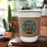 White, paper cup with Kraft Brown Coffee Sleeve displaying "your logo here", resting on a wood coaster, served with two cupcakes on a wooden table in a cafe