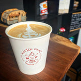 10 oz paper cup with red "Muster Point" logo, filled with milk coffee with floral foam pattern, on table, black bookshelf in background