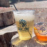 10 oz plastic cup and glass with Oaklore Distilling logo, filled with yellow and orange drinks on wooden stumps