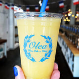 10 oz plastic cup with Olea logo and olive branches, yellow drink with blue straw, held by hand with blue nails in cafe