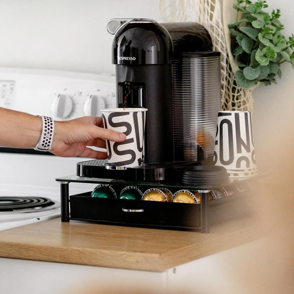 A person is using a Nespresso coffee machine, placing a 12 oz. custom-printed paper cup with a bold, modern design under the dispenser. A storage drawer below the machine holds various colored coffee pods. A watch is visible on the person's wrist. Nearby is a stove and a potted plant. The cups are printed by The Cup Store.