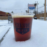 12 oz plastic cup with "Feed Store Beer Co" print, filled with dark beer with froth, standing in snow, street and fence in background