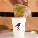 12 oz paper cup with burgundy "S" and bird logo, filled with milk coffee with froth, croissant being dipped into froth by hand, on table