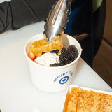 12 oz paper food container, with blue "Yogurt Club" logo, filled with ice cream and fruit by a tongs, on white table