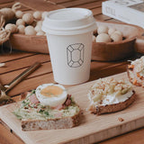 A 12 oz. custom-printed paper cup featuring a gem symbol stands on a wooden table next to an arrangement of open-faced sandwiches. The sandwiches include avocado toast with a soft-boiled egg and another topped with cream cheese and walnuts. A book and wooden beads lie in the background. The cups are printed by The Cup Store.