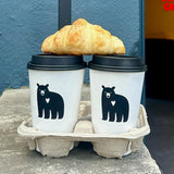 Two 16 oz double-walled paper cups with black dome lids and bear logo with white heart, in cardboard carrier with croissant, on stone ledge, blue wall