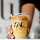 16 oz plastic cup with black "Savage Coffees" logo, filled with iced milk coffee, being placed on white table by hand