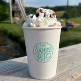 16 oz paper cup with blue "The Sweet Spot" logo, filled with milk coffee, whipped cream, chocolate syrup, clear straw, on wooden table, park in background