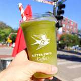 20 oz plastic cup with bird logo and "Bitter Pops Coffee" text, filled with green drink, sip lid and red-white straw, held by hand with street background