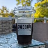 20 oz plastic cup with sip lid, white "Coldbruh" logo, filled with dark coffee and froth, standing on wet table on patio