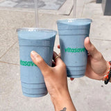 Two 24 oz plastic cups with green "Vitasave" print, filled with blue drinks, flat lids and clear straws, held by hands over tile floor