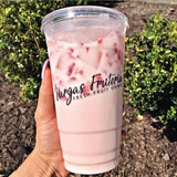 32 oz plastic cup with black "Vargas Fruteria" logo, flat lid, filled with strawberry milk drink with strawberries and ice, held by hand, bushes in background
