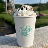 A cup of dessert featured in a 16 oz custom-printed paper cup with The Sweet Spot logo, topped with whipped cream and chocolate syrup, sits on a wooden ledge with a blurred outdoor setting in the background. Cups printed by The Cup Store.