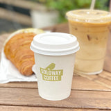 8 oz paper cup with white dome lid, green "Soloway Coffee" logo with bird, next to plastic iced coffee cup and croissant on wooden table, park in background