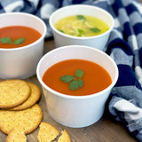 Three 12 oz paper food containers, filled with tomato and chicken noodle soup, on a wooden table next to a blanket and crackers