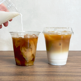 Two 12 oz plastic cups with coffee, milk, and ice, left cup being poured into by hand, right cup with sip lid, on wooden table with white wall