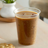 16 oz plastic cup filled with iced coffee on white table, cookie on napkin beside it, potted plant on wooden tray in background