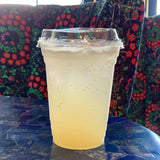 16 oz blank plastic cup with sip lid, filled with sparkling lemonade, standing on blue table with decorative flower pillows in background